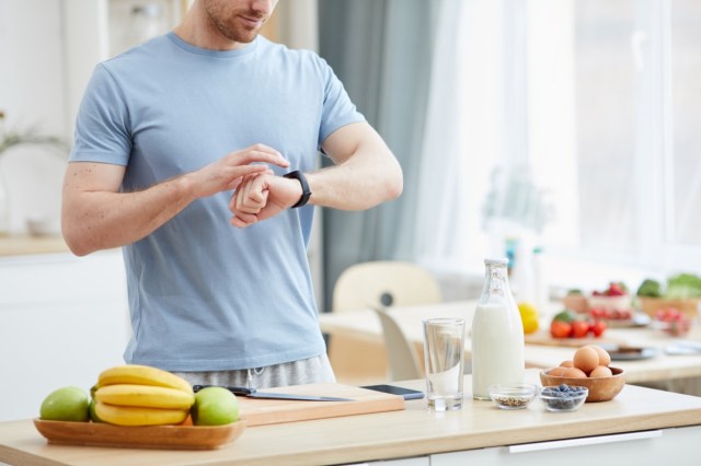 A man standing near the kitchen table with food and checking his watch.