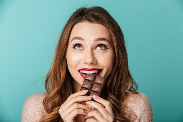 Portrait of a delighted brown haired woman with bright makeup eating chocolate bar